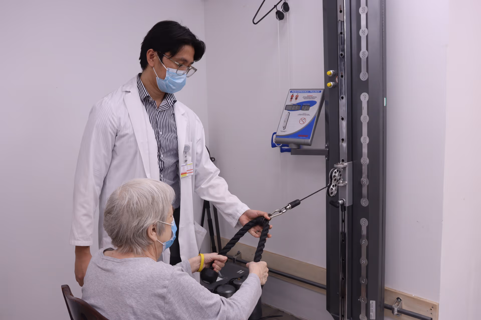 A healthcare professional wearing a white coat and face mask assists an elderly woman, also wearing a face mask, in using a resistance exercise machine in a clinical setting.