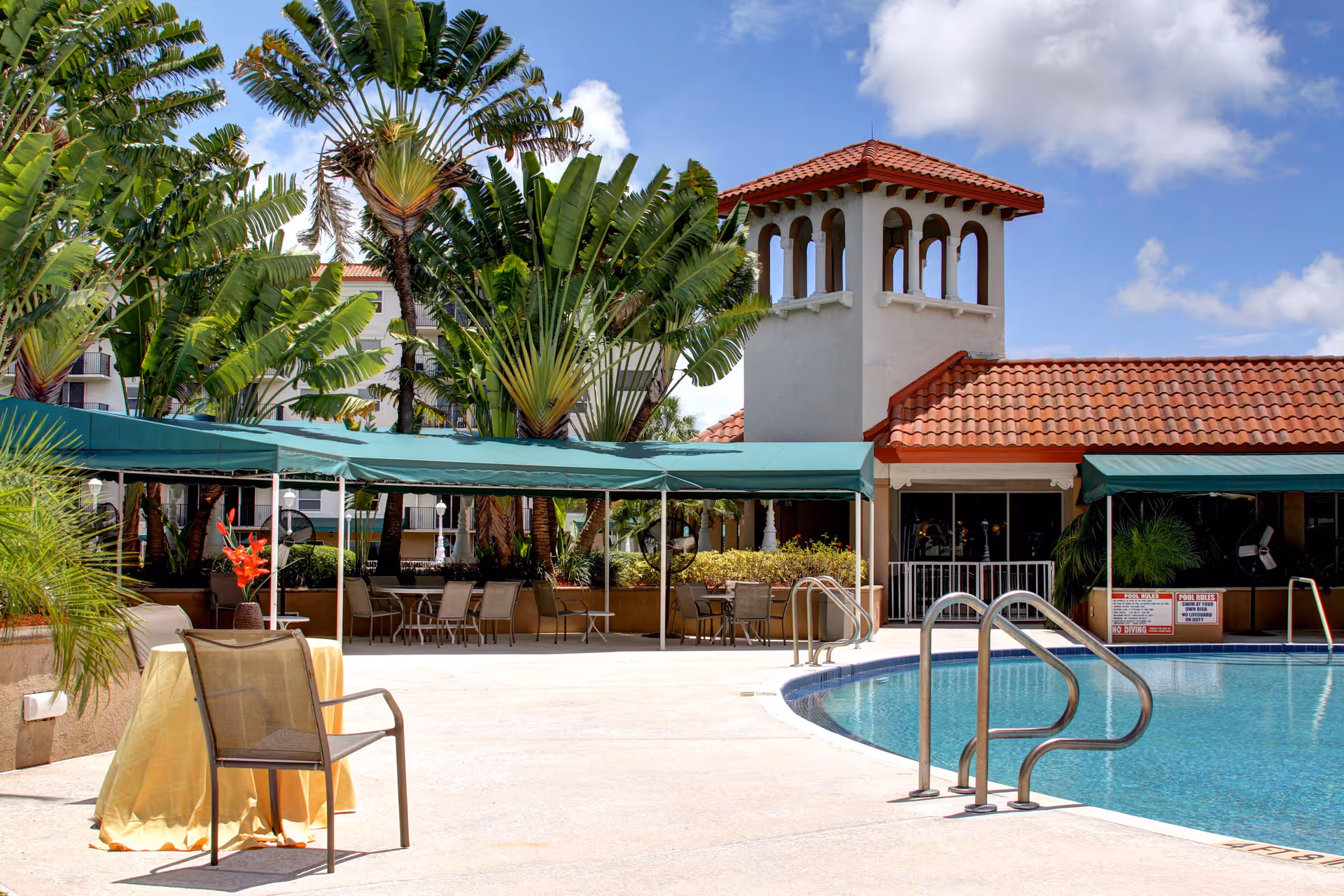 Outdoor swimming pool with metal handrails, patio seating, palm trees, and a red-tile roof building.
