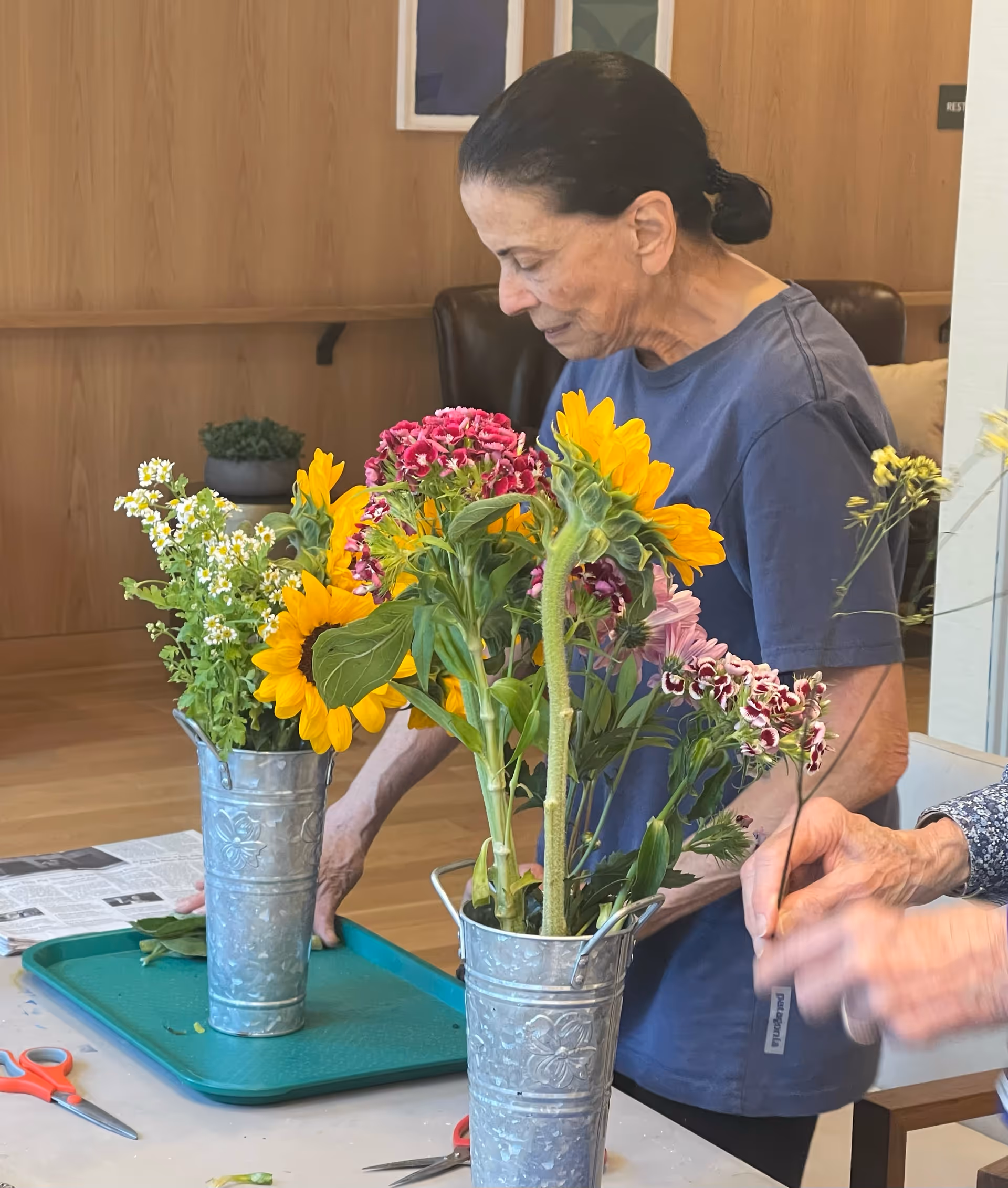An elderly woman arranging flowers in metal vases on a table indoors, with another person's hands also visible arranging flowers. The setting appears to be a cozy room with wooden walls and a leather chair in the background.