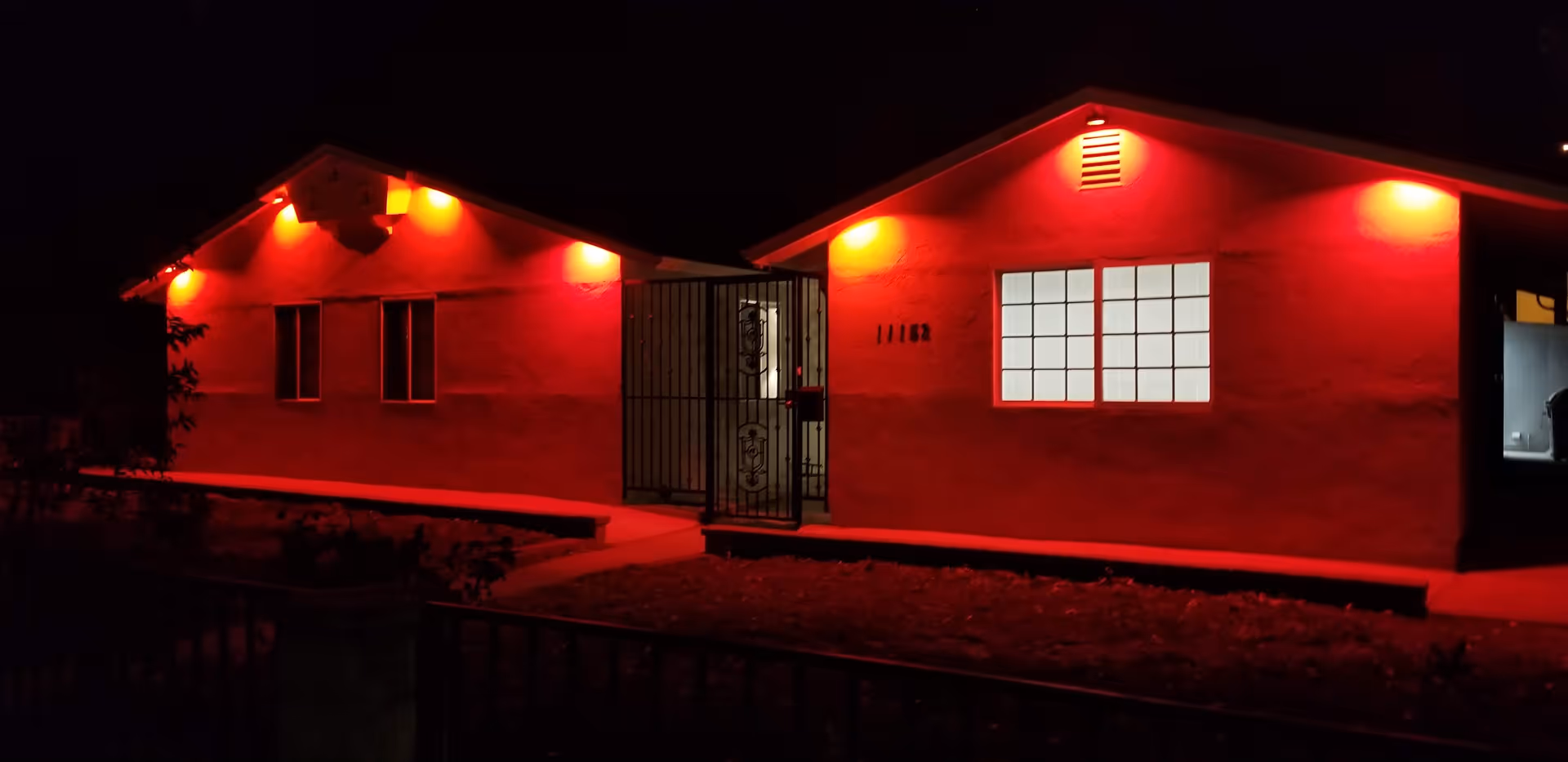 Exterior view of a single-story building at night illuminated by red lights above the windows and entrance. The building has a gated entrance and a window with white light shining through. The surrounding area is dark with some visible plants and a fence in the foreground.