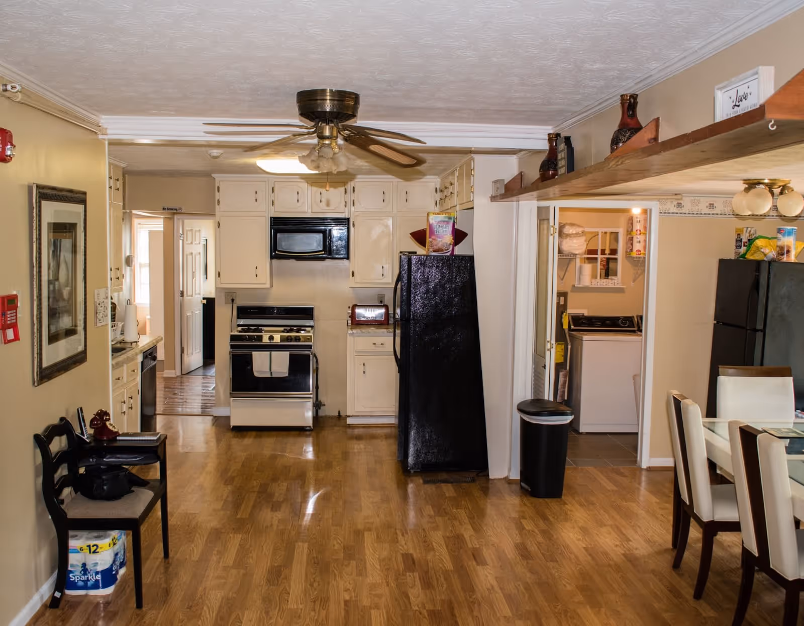 Interior view of a kitchen and dining area in a personal care home. The kitchen features white cabinets, a black refrigerator, a stove with an oven, a microwave, and a ceiling fan with lights. To the right, there is a dining table with white cushioned chairs. A doorway leads to a laundry room with a washing machine and shelves stocked with paper towels and other supplies. The floor is wooden, and the walls are painted beige. Various decorative items and kitchen essentials are visible on shelves and counters.
