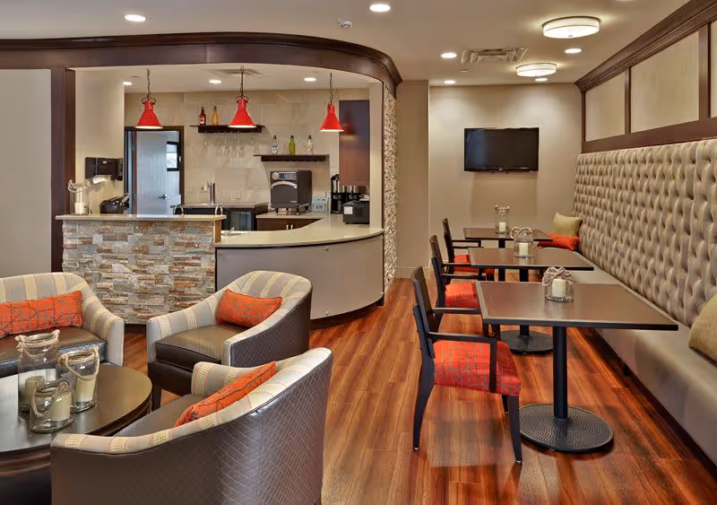 A modern communal dining and lounge area with chairs, small tables, a tufted banquette, a service counter with red pendant lights, and a wall-mounted TV.