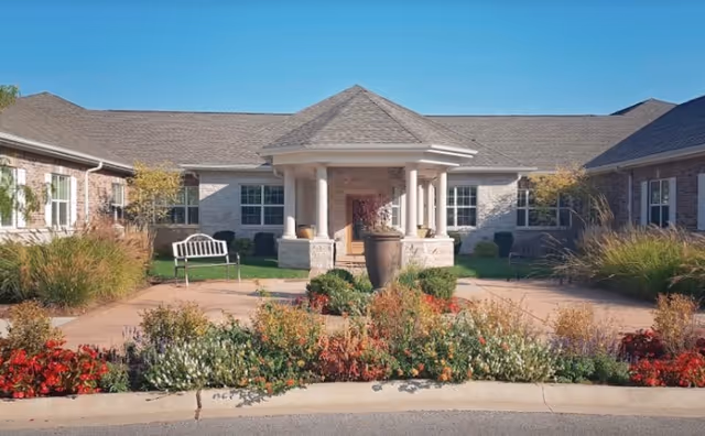 Front exterior view of Churchill Place Memory Care facility showing a single-story building with a covered entrance supported by columns, surrounded by landscaped gardens with colorful flowers, shrubs, and a bench on the left side.