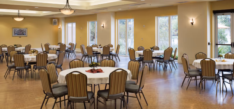 A spacious dining room with multiple round tables covered with white tablecloths and surrounded by striped cushioned chairs. The room has large windows letting in natural light and yellow walls with wall sconces and ceiling lights.
