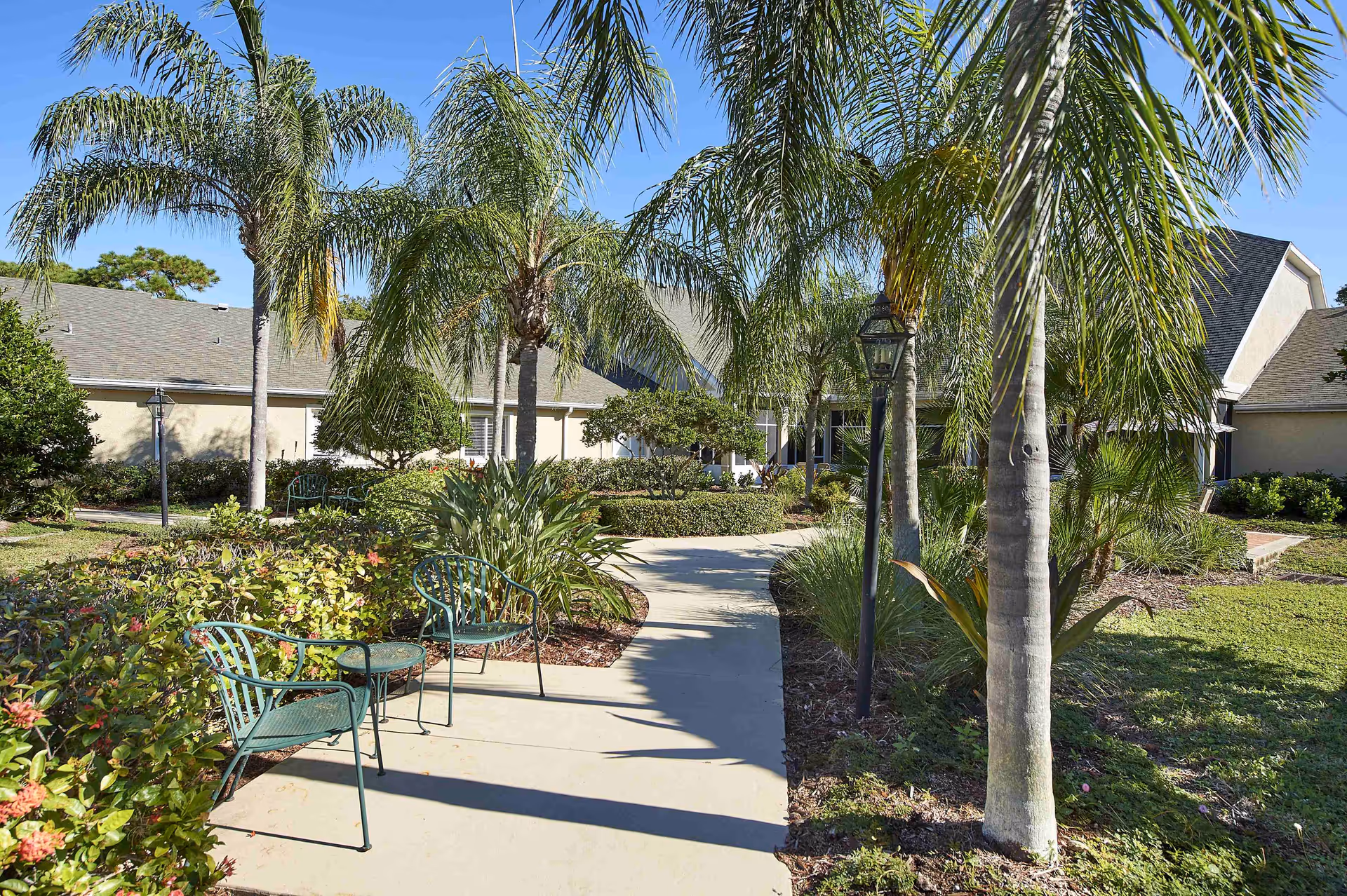 Outdoor pathway lined with palm trees and green plants leading to a building with a gray roof. Two green metal chairs and a small table are placed along the walkway under the shade of the trees.