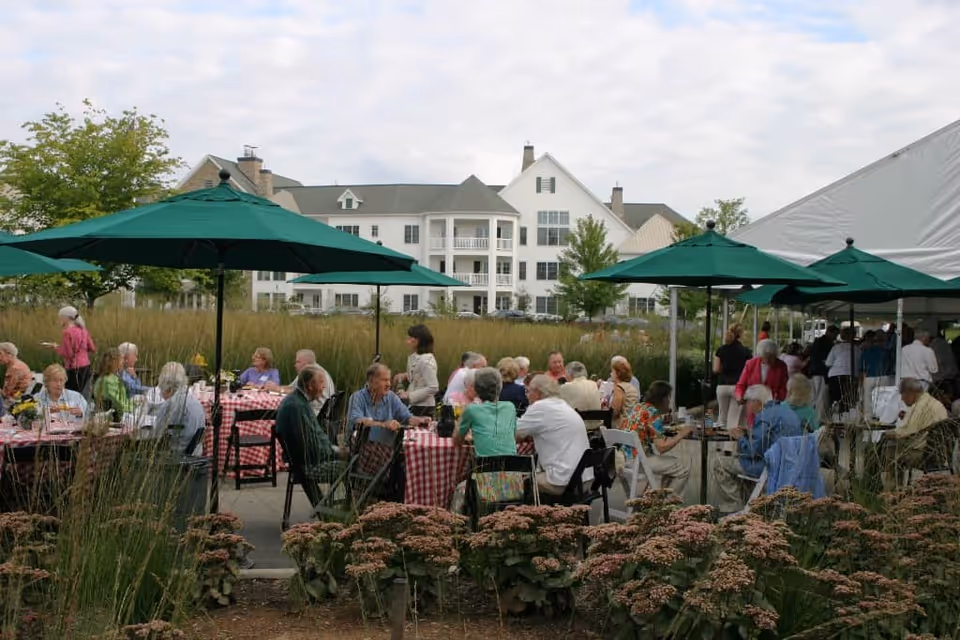 Outdoor gathering at South Franklin Circle, a Judson Senior Living Community, with senior residents seated at tables covered with red and white checkered tablecloths under green umbrellas. The background shows a large white multi-story building and some greenery.