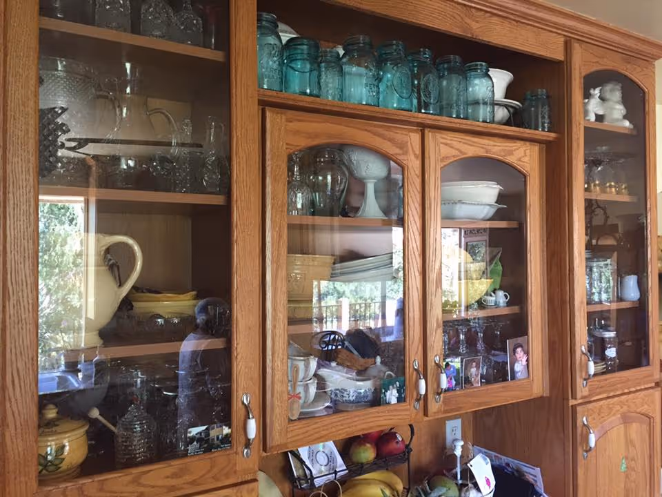 Wooden kitchen cabinets with glass doors displaying various glassware, dishes, jars, and decorative items. Below the cabinets, there is a countertop with fruit and other kitchen items visible.