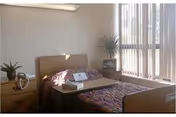 Small nursing home bedroom with a single bed, bedside tables, potted plants and vertical blinds letting in daylight.