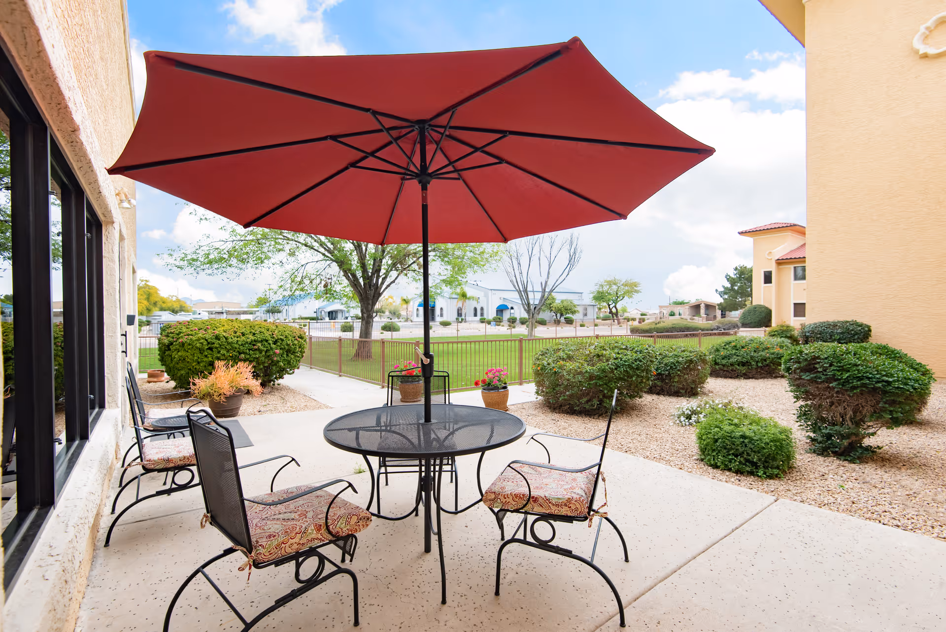 Outdoor patio area with a round metal table and four cushioned chairs under a large red umbrella. The patio is surrounded by landscaped bushes and plants, with a grassy area and trees visible beyond a fence. The sky is partly cloudy.