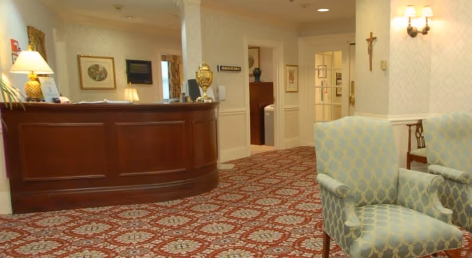Reception area with a wooden front desk, two upholstered armchairs with a blue and gold pattern, patterned carpet, wall lamps, framed artwork, and a crucifix on the wall.