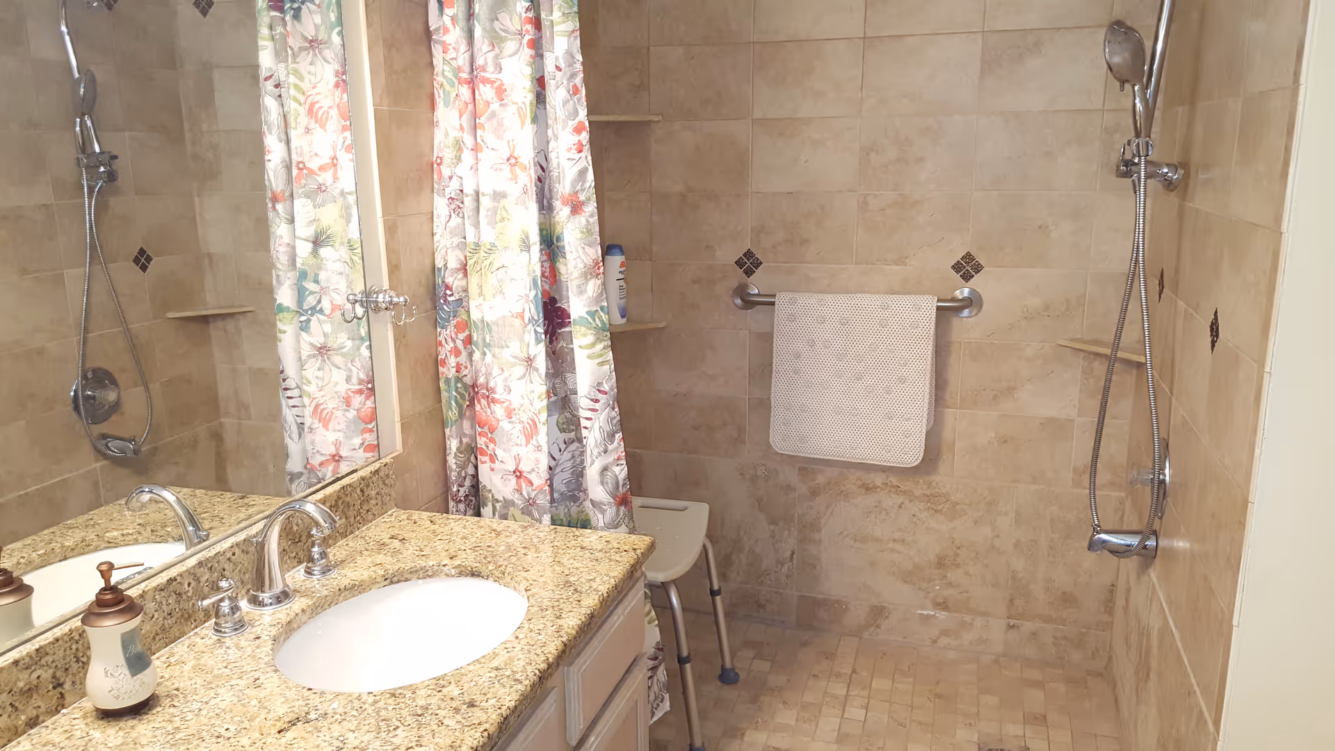 Bathroom with beige tiled walls and floor, featuring a walk-in shower with a handheld showerhead, a white shower chair, and a grab bar with a white towel hanging on it. There is a granite countertop with a sink and a floral patterned shower curtain reflected in a large mirror above the sink.
