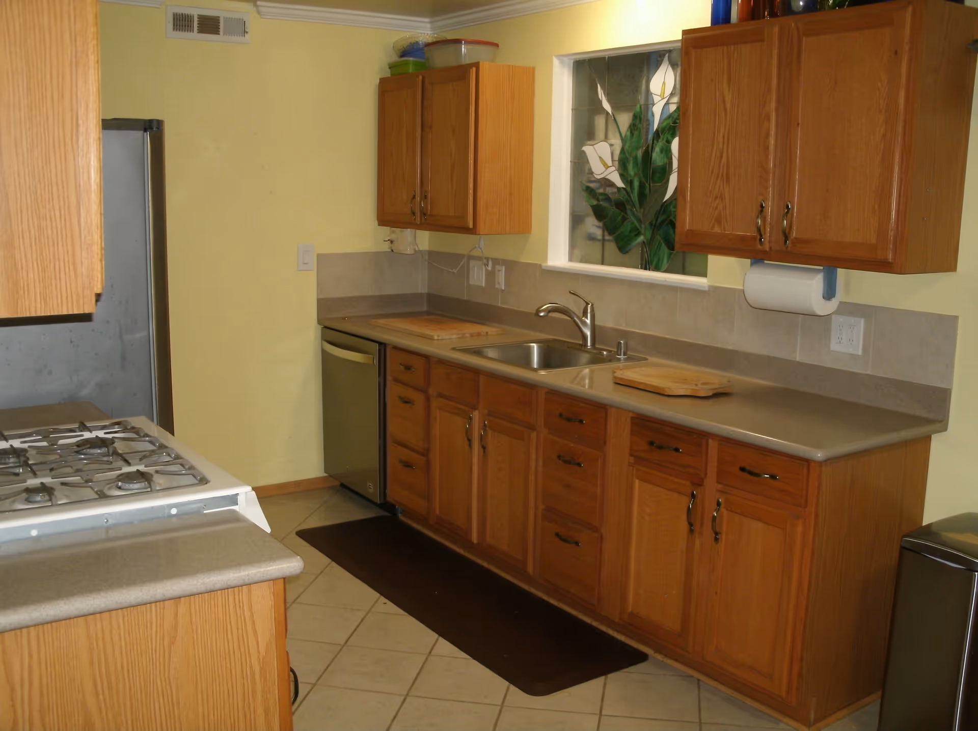 A kitchen with wooden cabinets, a stainless steel sink, a dishwasher, a gas stove, and a refrigerator. The walls are painted yellow, and there is a decorative stained glass window above the sink. Two wooden cutting boards are placed on the countertop.