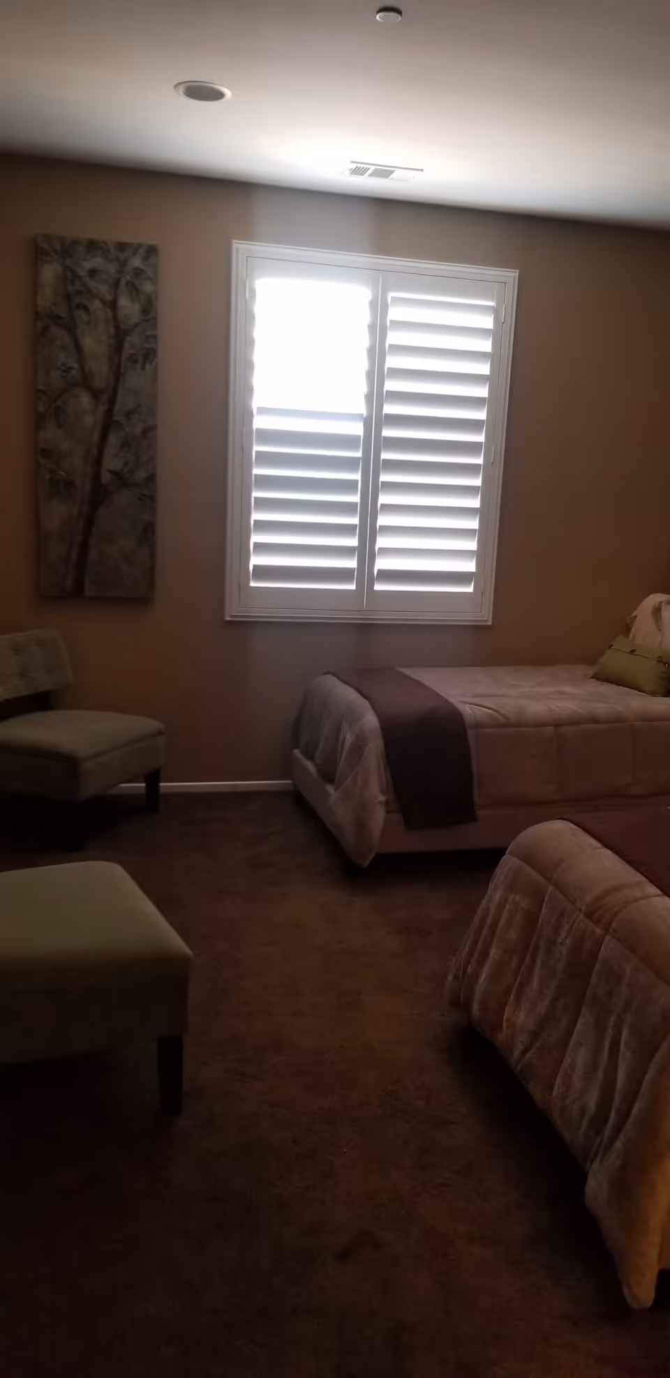 A bedroom with two twin beds covered in beige and brown bedding. There is a window with white plantation shutters letting in light. A cushioned chair and ottoman are positioned near the left wall, which has a vertical decorative painting of a tree.