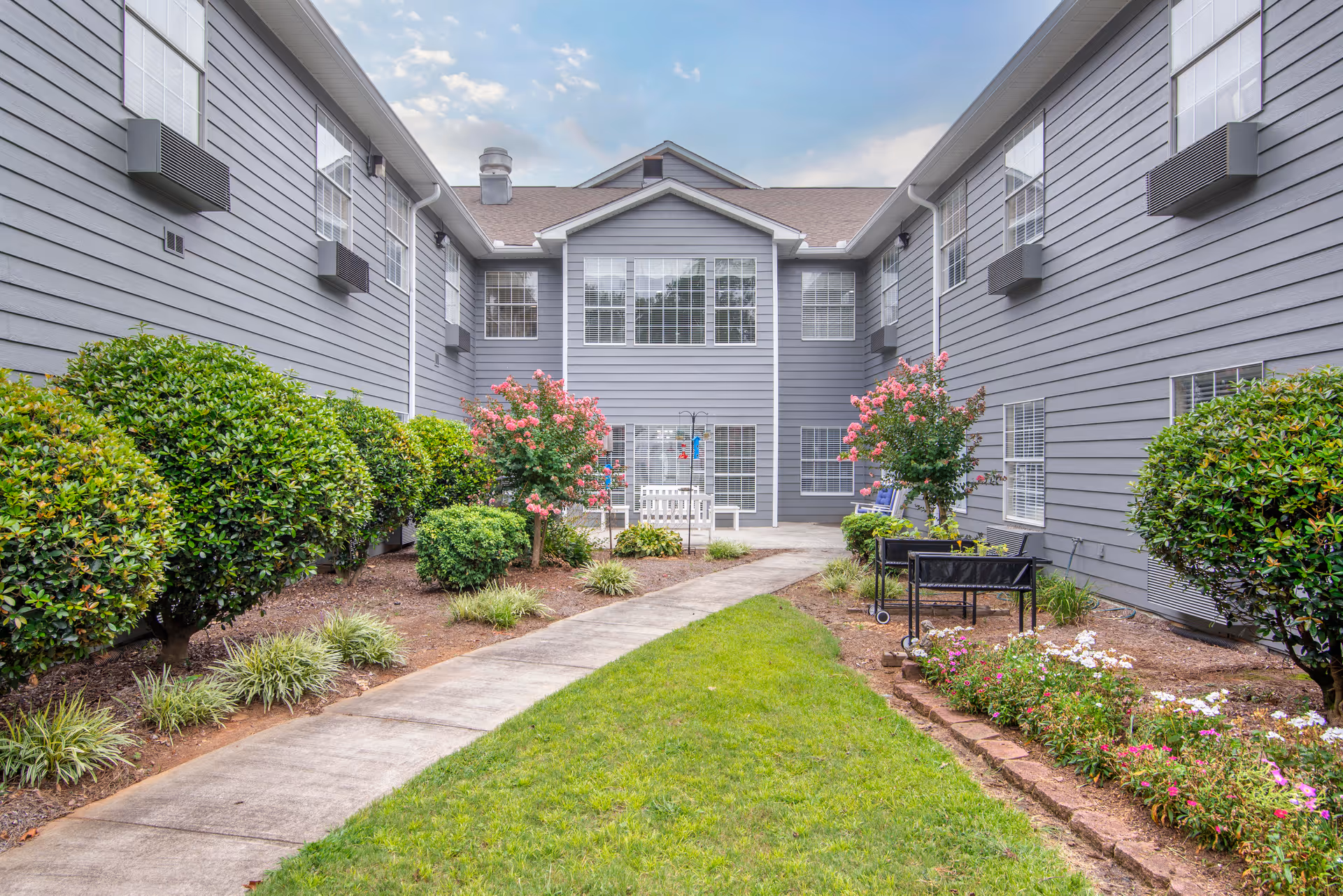 A landscaped courtyard area of a senior living facility with a concrete pathway, green grass, trimmed bushes, flowering plants, and two pink flowering trees. The courtyard is surrounded by a two-story gray building with multiple windows.