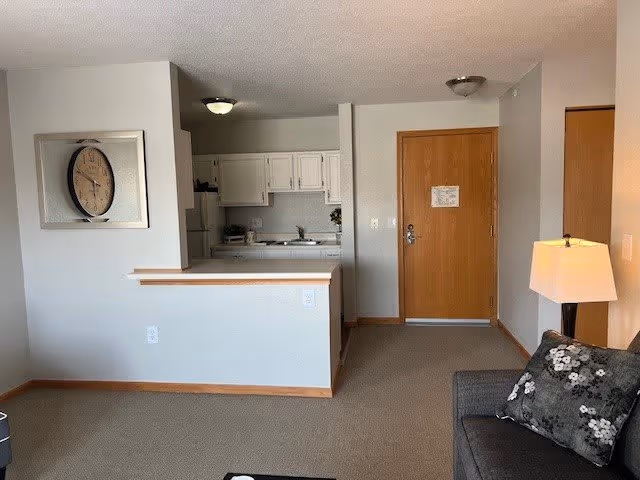 Interior view of a senior living facility apartment showing a small kitchen area with white cabinets and appliances, a wooden entrance door, a gray sofa with a floral pillow, a floor lamp, and a wall clock mounted on a light gray wall.