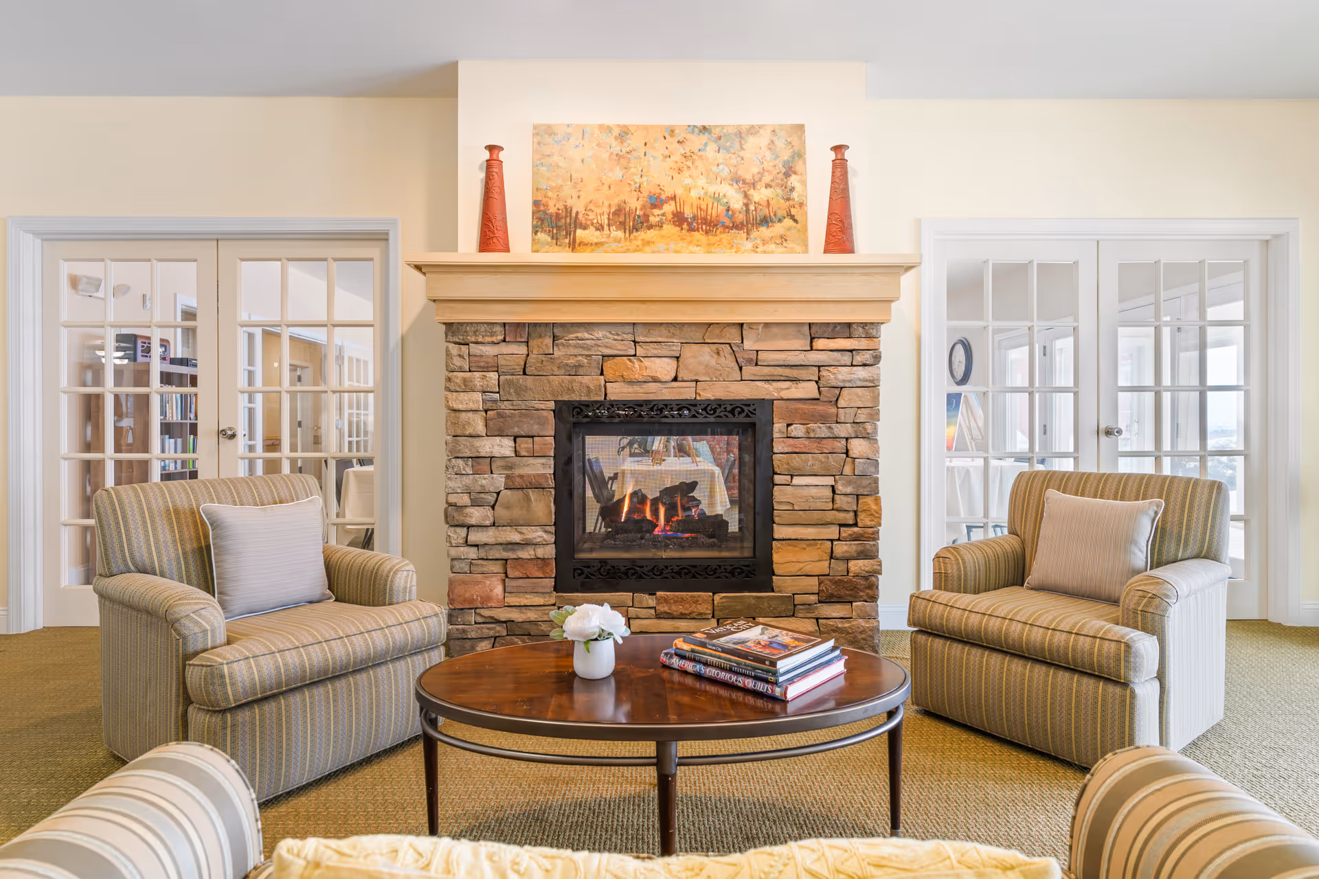 Cozy seating area with a stone fireplace flanked by two upholstered armchairs and a round coffee table with books and a small plant.