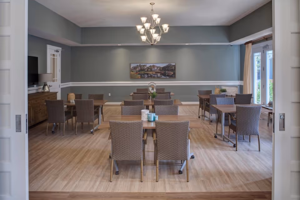A dining room with several wooden tables and wicker chairs arranged neatly on a light wood floor. The walls are painted gray with white trim, and a chandelier with multiple lights hangs from the ceiling. A large painting is centered on the far wall, and there are large windows with curtains on the right side letting in natural light.