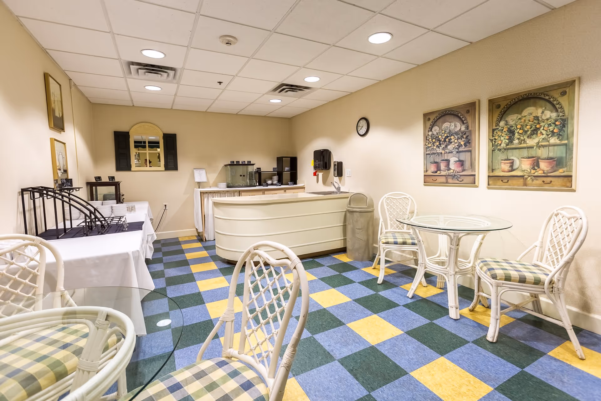 A small dining area with checkered blue, green, and yellow flooring, featuring glass-top tables with white wicker chairs that have plaid cushions. The room has a counter with a coffee station, a trash can, wall-mounted soap and paper towel dispensers, and decorative paintings of potted plants on the wall. The ceiling has recessed lighting and there are framed pictures and a mirror on the opposite wall.