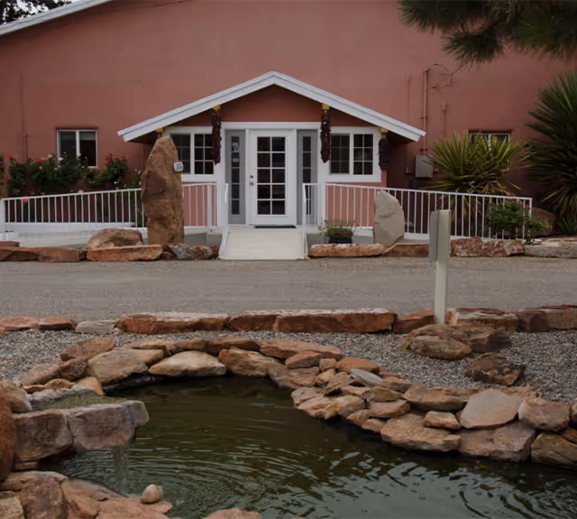 Front entrance of a pink stucco assisted living building with white double doors, a ramp, and a rock-lined pond in the foreground.