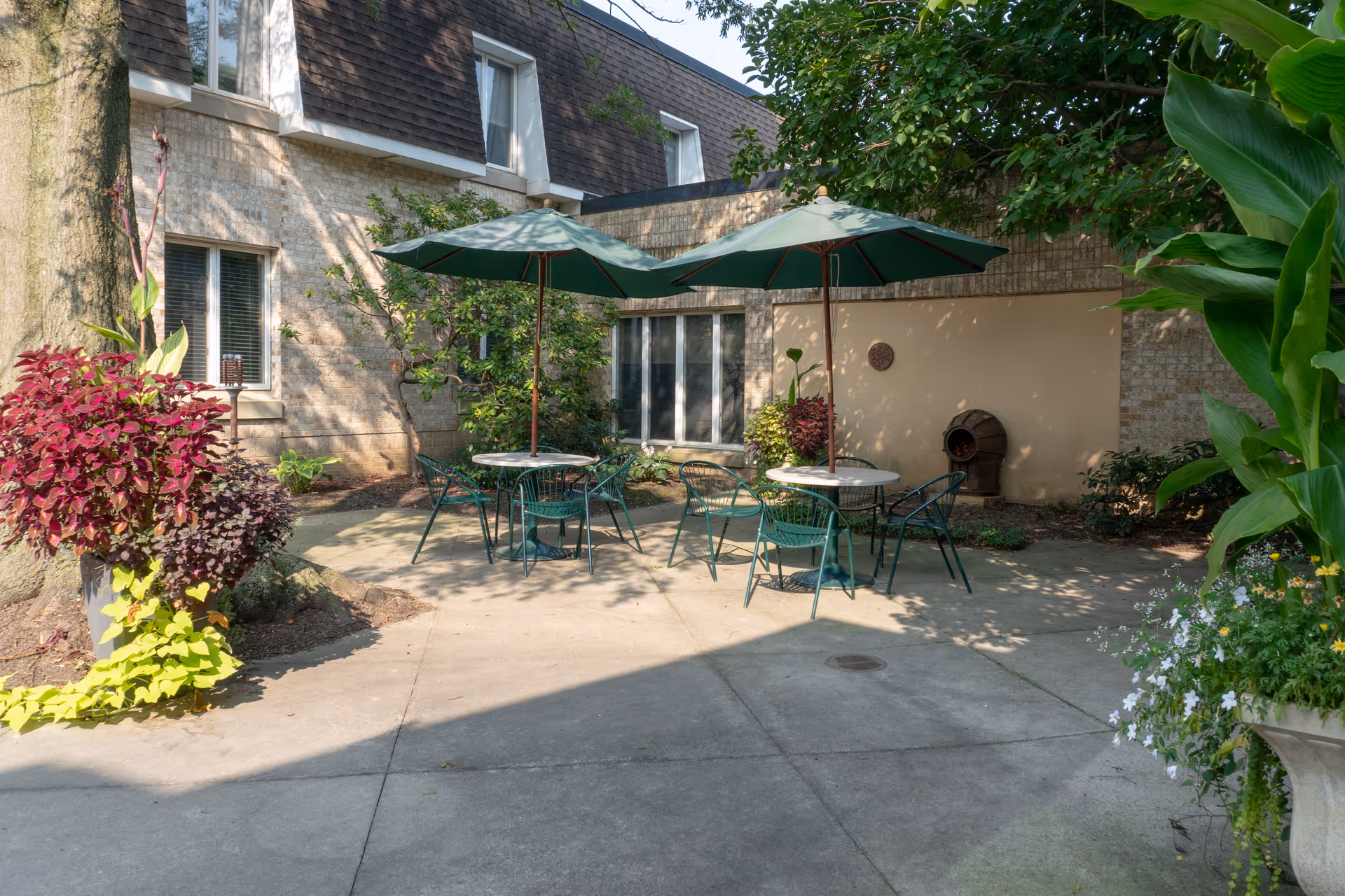 Outdoor patio area with two round tables, each shaded by a green umbrella, surrounded by green metal chairs. The patio is adjacent to a building with windows and surrounded by lush plants and trees.