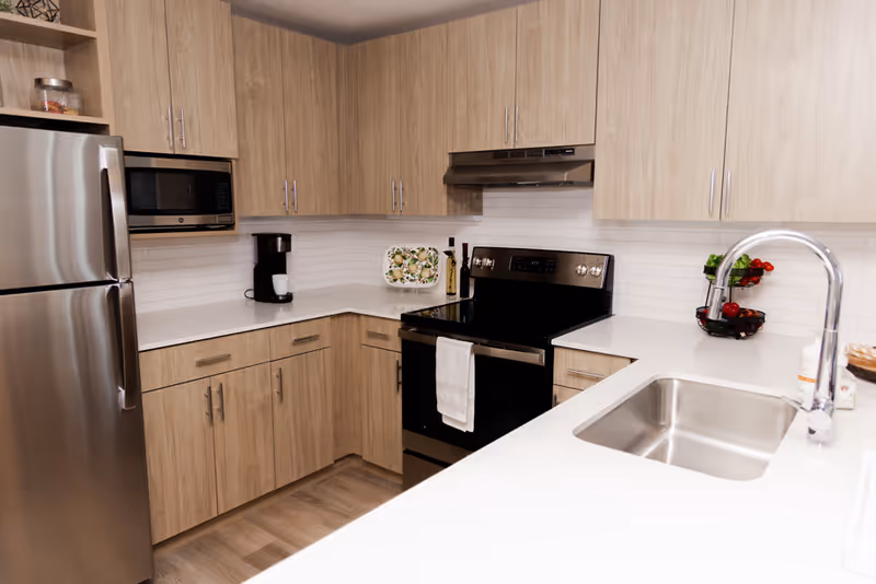 Modern kitchen with light wood cabinets, stainless steel refrigerator, microwave, black stove with a white towel hanging on the handle, white countertops, a coffee maker, a two-tier fruit basket with vegetables, and a stainless steel sink with a curved faucet.