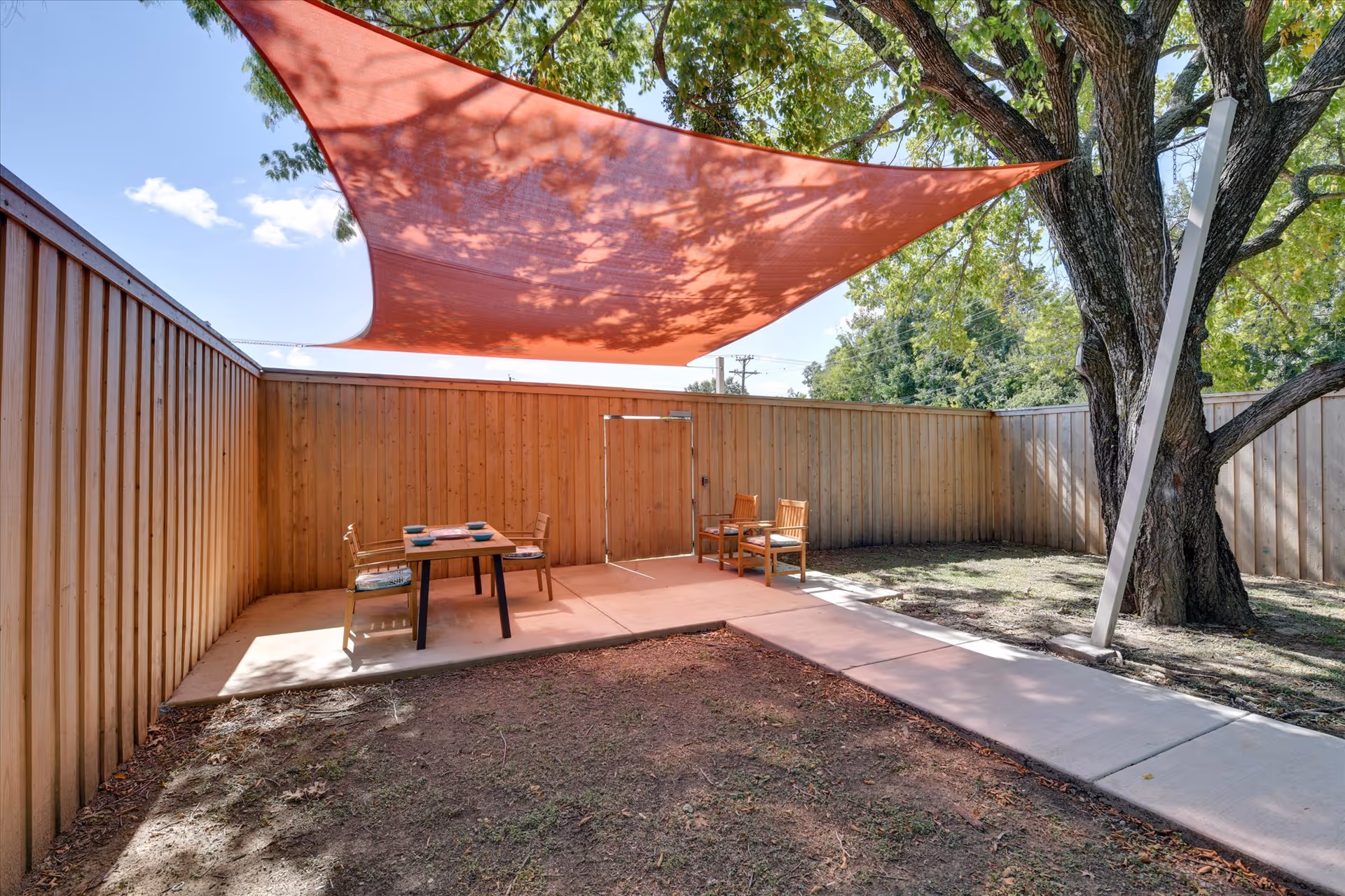Outdoor patio area with a wooden fence, a large tree, and a red triangular shade sail overhead. There are two sets of wooden chairs and tables on a concrete slab, with a concrete walkway leading to a gate in the fence.