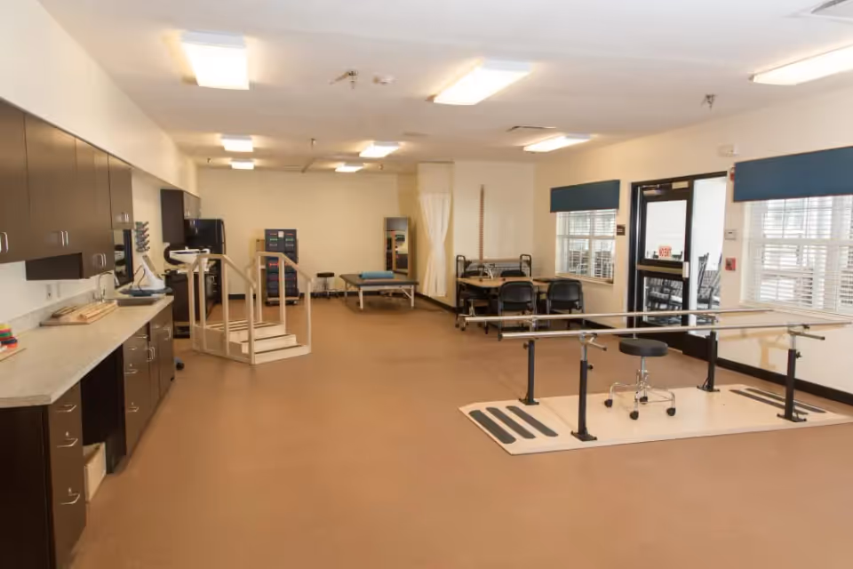 A spacious rehabilitation room with parallel walking bars, exercise stairs, treatment tables, cabinets, and seating.