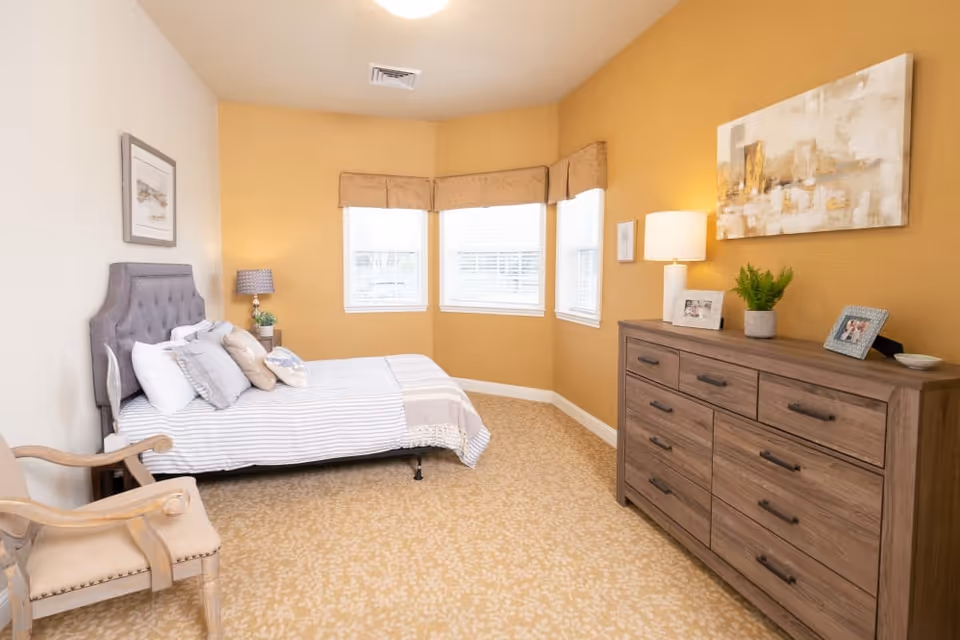 A cozy bedroom with a bed, upholstered headboard, armchair, wooden dresser with lamp and decor, and bay windows on warm yellow walls.
