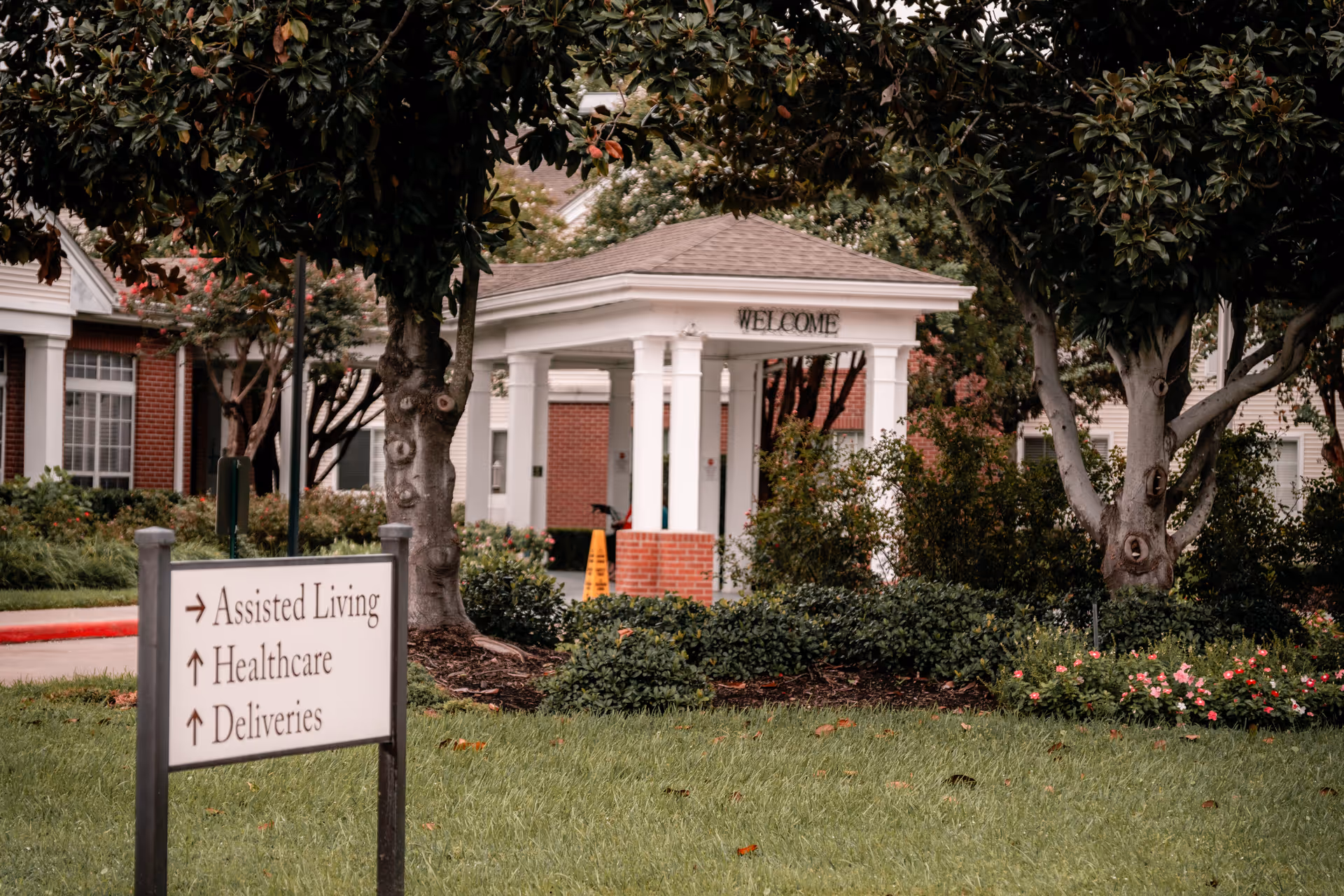 Outdoor view of a senior living facility entrance with a white pavilion that has a 'WELCOME' sign on it. There are trees, bushes, and flowers surrounding the area. A sign in the foreground directs to Assisted Living, Healthcare, and Deliveries.