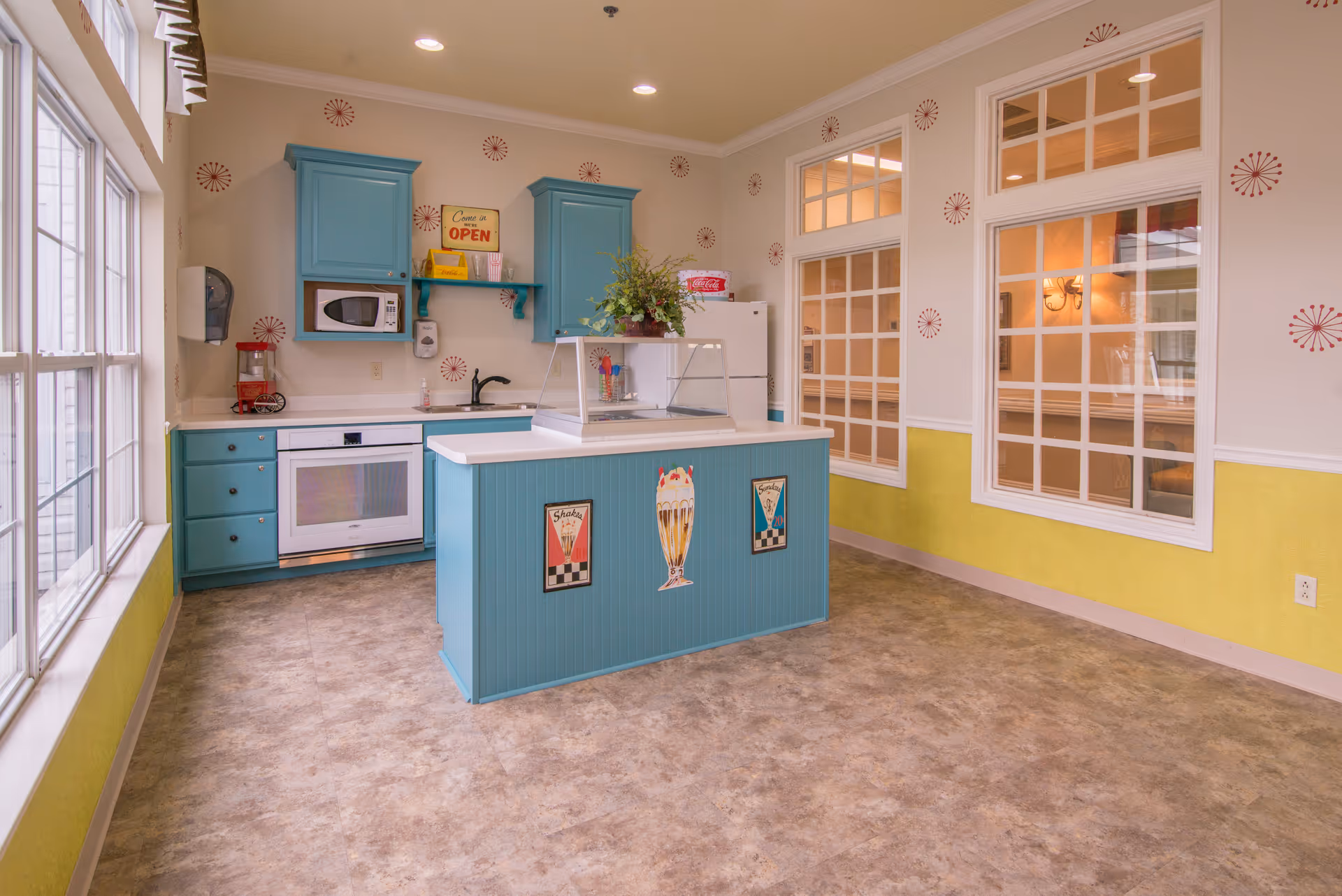 A bright kitchen area with teal cabinets and a white countertop island decorated with vintage-style milkshake signs. The kitchen includes a microwave, oven, sink, and refrigerator. The walls are decorated with red starburst patterns and have large windows and glass-paneled interior windows letting in natural light. A small plant sits on the island countertop.