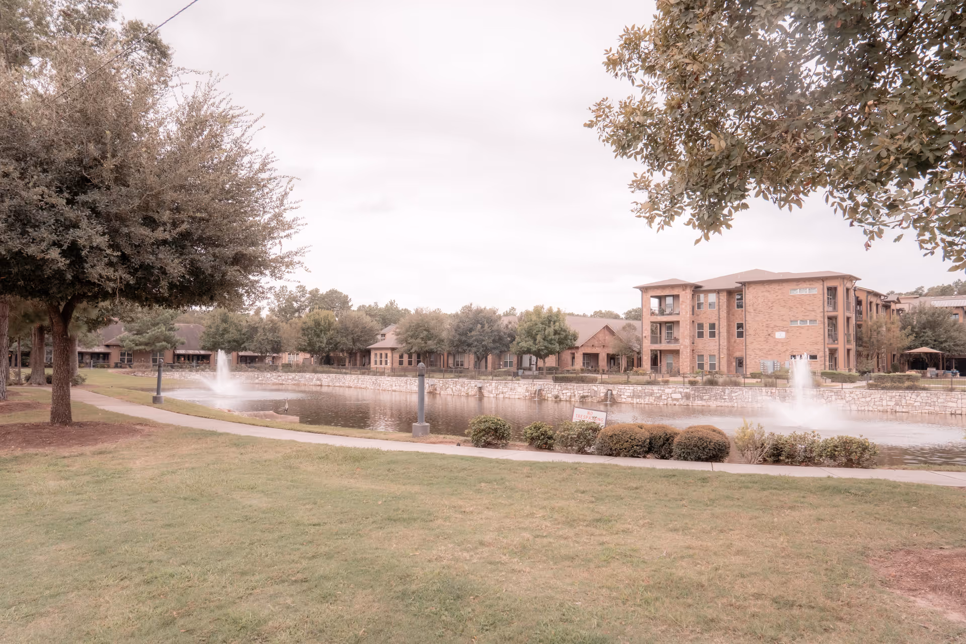 A landscaped outdoor area of an assisted living facility featuring a pond with two water fountains, surrounded by a stone wall and walking paths. There are trees, bushes, and a grassy lawn in the foreground, with multi-story brick buildings in the background under a cloudy sky.