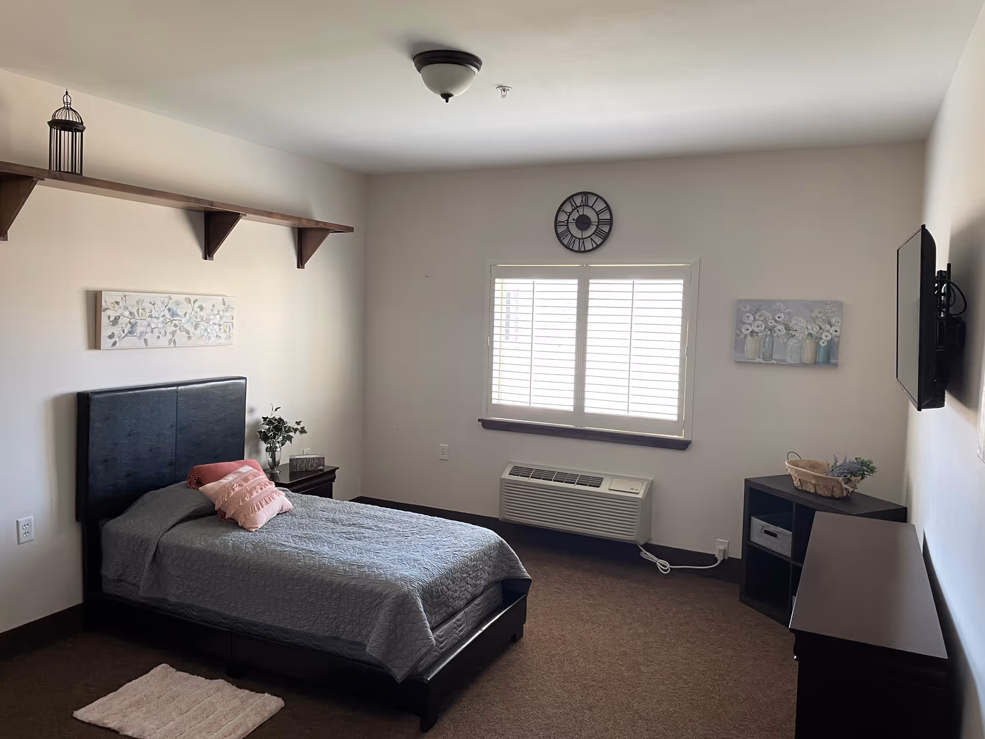 A simple bedroom with a single bed covered in a gray quilt and pink pillows. The room has a window with white shutters, a wall clock above the window, a mounted TV on the right wall, a small nightstand with a plant and decor, a corner shelf unit with a basket and plant, and a wooden shelf mounted high on the left wall. The walls are light-colored and the floor is carpeted.