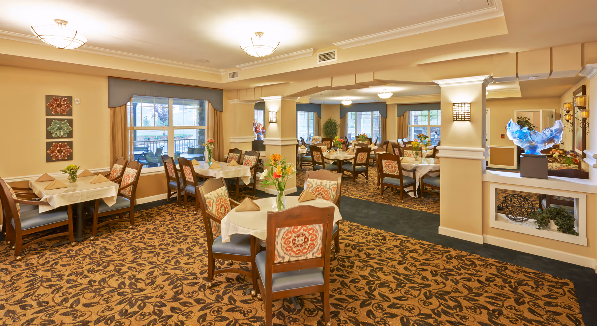 A spacious dining room with multiple tables covered in white tablecloths and set with napkins and flower centerpieces. The room has patterned carpet, large windows with curtains, and warm lighting fixtures on the ceiling and walls. Decorative art pieces and plants are placed around the room, creating a welcoming atmosphere.
