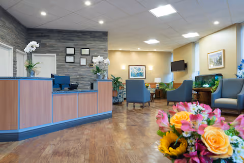 Reception area of a senior living facility with a wooden front desk, computer, and potted plants. The room has wooden flooring, recessed ceiling lights, and a seating area with blue armchairs and a fish tank. A colorful bouquet of flowers is in the foreground.