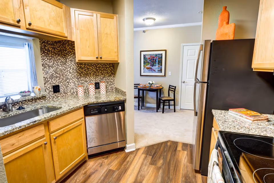 Bright kitchen with wooden cabinets, granite countertops, stainless appliances, and a small dining table visible through a doorway.