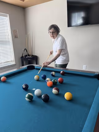 An elderly woman playing pool in a room with a blue pool table, a wall-mounted TV, and a window with blinds letting in natural light.