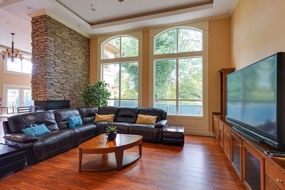 A spacious living room with a large black leather sectional sofa adorned with blue and yellow pillows, a wooden coffee table with a small plant, a stone fireplace, large arched windows letting in natural light, and a flat-screen TV on a wooden cabinet.