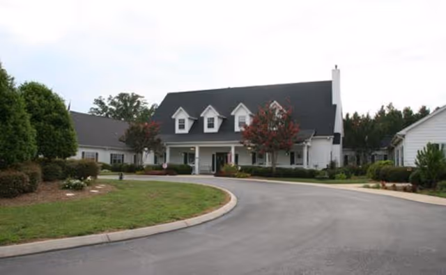 Front view of a white two-story building with dormer windows, a covered entrance, curved driveway, and landscaped grounds.