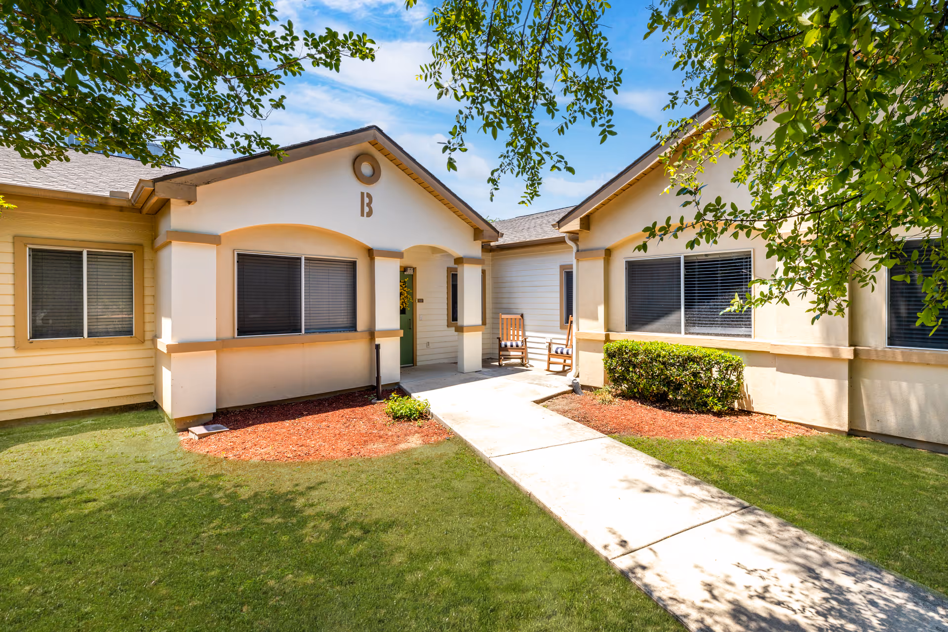 Exterior view of a single-story senior living facility building with beige walls and a green door. There is a concrete walkway leading to the entrance, flanked by green grass, mulch beds, and bushes. Two wooden rocking chairs with cushions are placed near the entrance under a covered porch. Trees with green leaves frame the top of the image under a blue sky with some clouds.