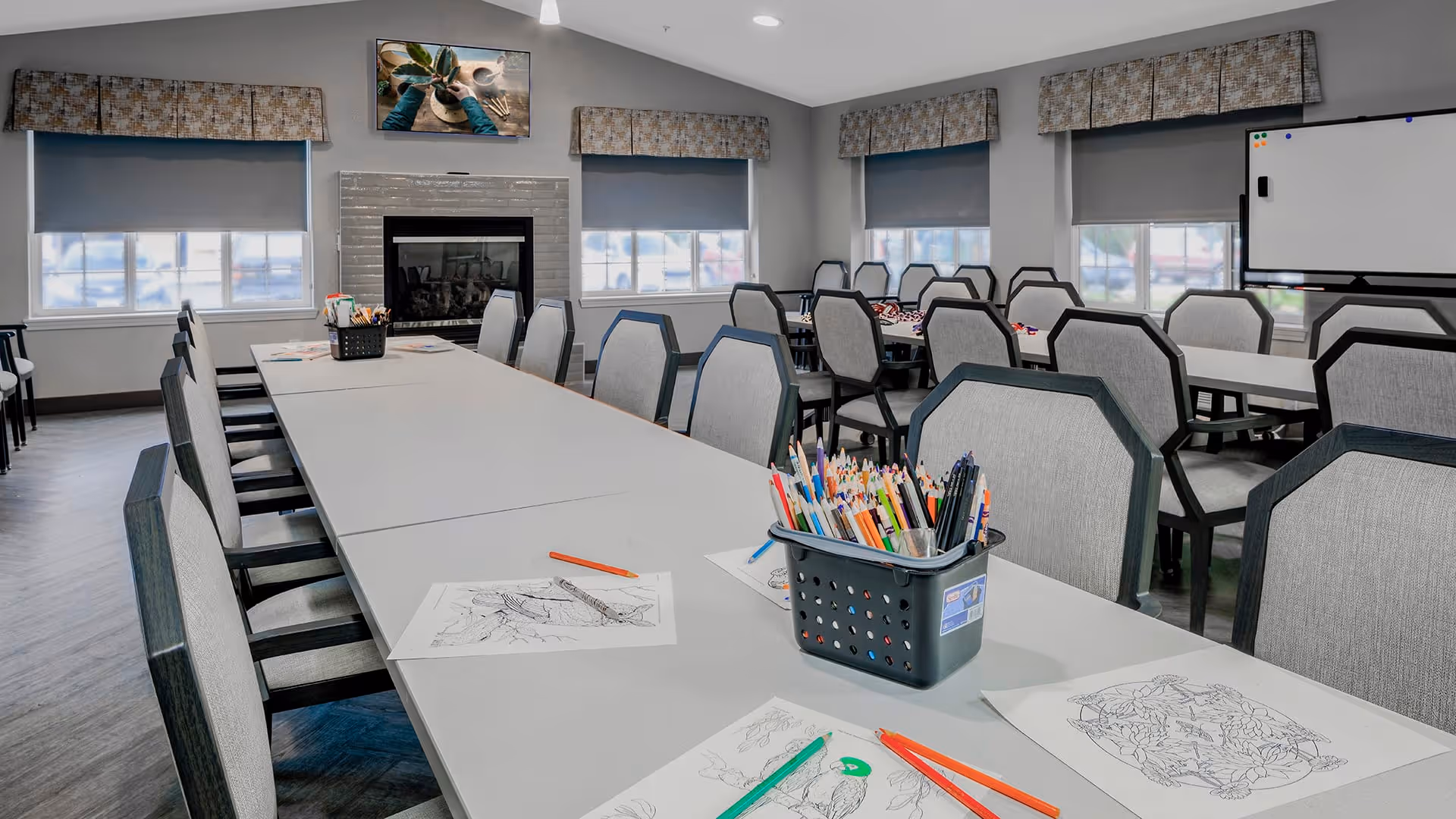 A well-lit activity room with long tables and multiple chairs arranged around them. On the tables are coloring sheets and containers filled with colored pencils and markers. The room has large windows with blinds, a fireplace, and a whiteboard in the background.