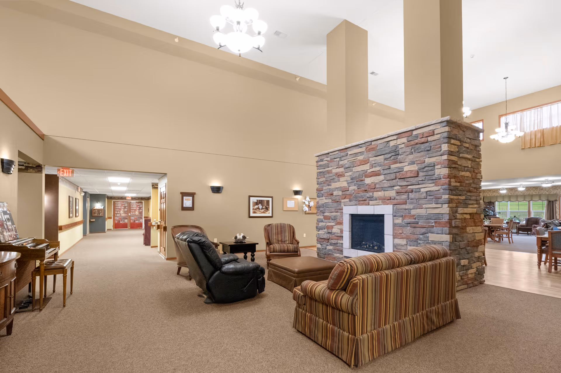 A spacious senior living facility common area with a stone fireplace in the center. Surrounding the fireplace are comfortable seating options including a striped sofa, a leather recliner, and an armchair with an ottoman. The room has beige walls, carpeted floors, and is well-lit with ceiling lights and wall sconces. In the background, there is a hallway and a dining area with tables and chairs.