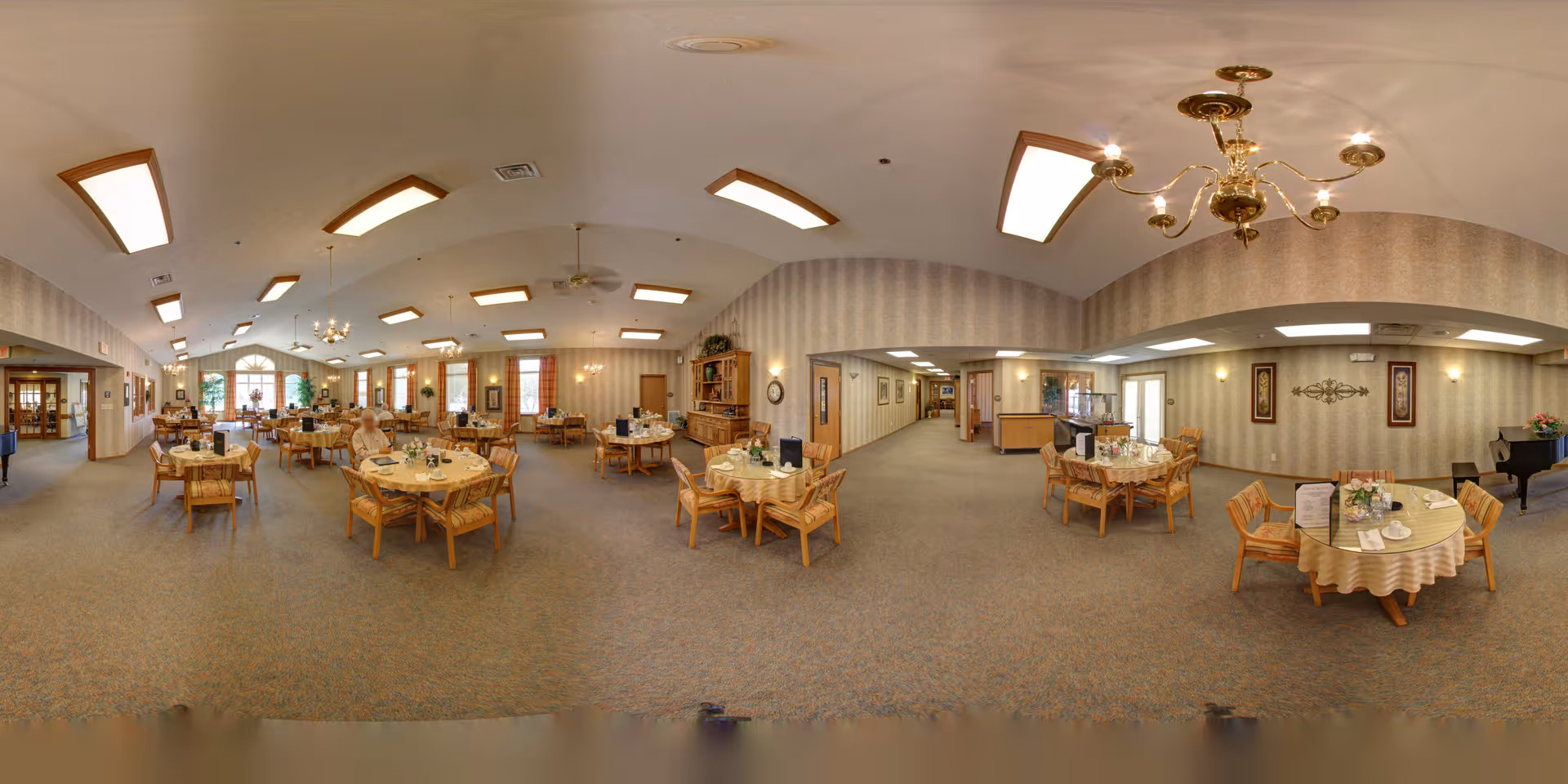 A spacious dining room in a senior living facility with multiple round tables covered with tablecloths and set for meals. The room has beige patterned wallpaper, carpeted floors, and several chandeliers and ceiling lights. There are large windows with curtains letting in natural light, and a piano is visible in the corner. Some tables have menus and floral centerpieces.