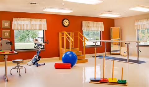 A physical therapy or rehabilitation room with exercise equipment including a stationary bike, a blue exercise ball, a red foam roller, parallel bars for walking practice, and a set of colorful balance stepping stones. The room has large windows with beige curtains, a clock on a red accent wall, and a wooden staircase for therapy exercises.