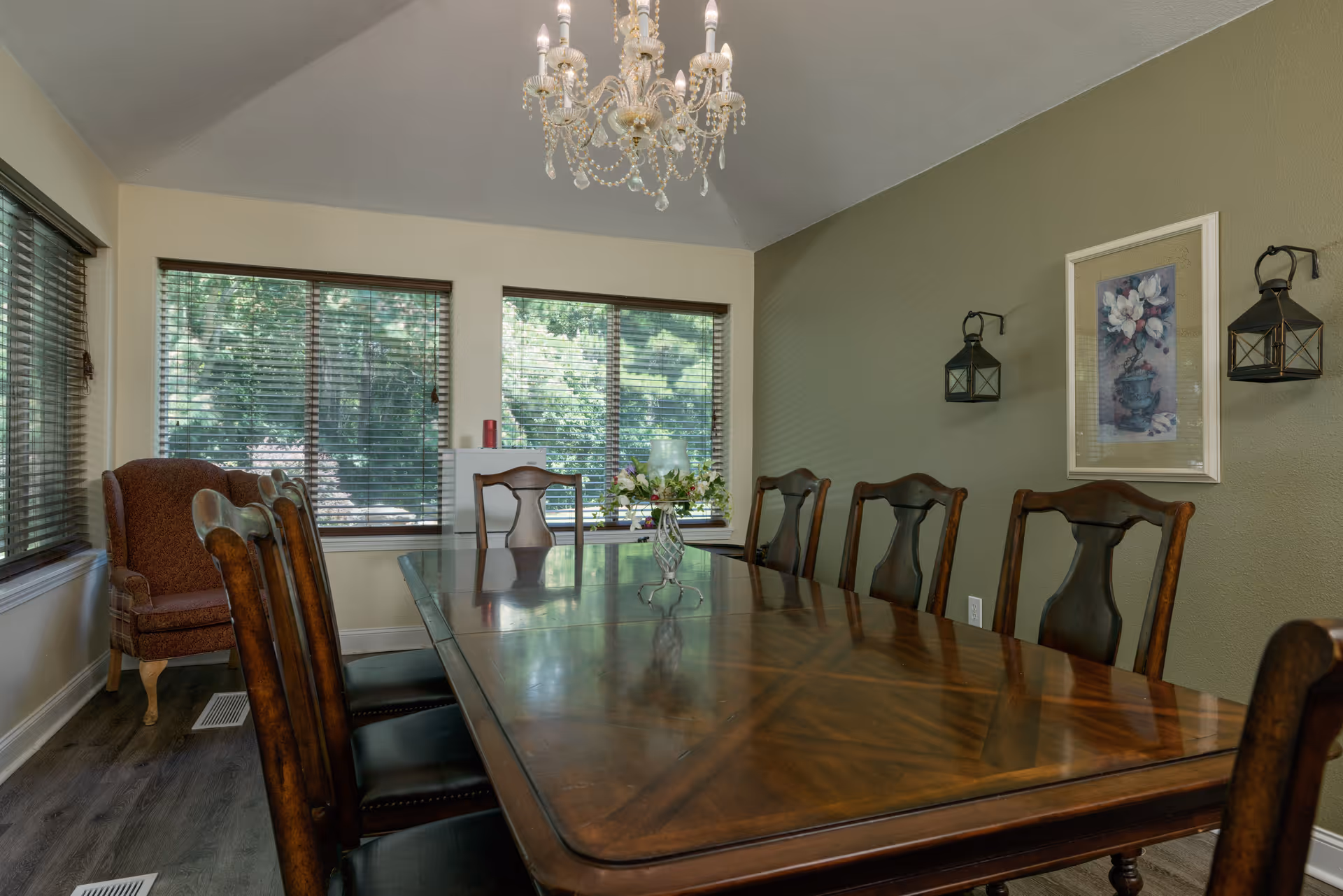 Formal dining room with a long polished wooden table surrounded by chairs, a chandelier overhead, large windows with blinds, and wall decor.