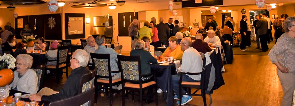 A large group of older adults seated at tables and standing while socializing in a decorated communal dining/activity room.