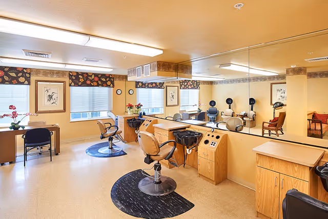 Interior of a hair salon area in a senior living facility with salon chairs, hair washing stations, large mirrors on the wall, and a desk with a chair near the window. The room has beige walls, floral valances on the windows, and decorative framed artwork.