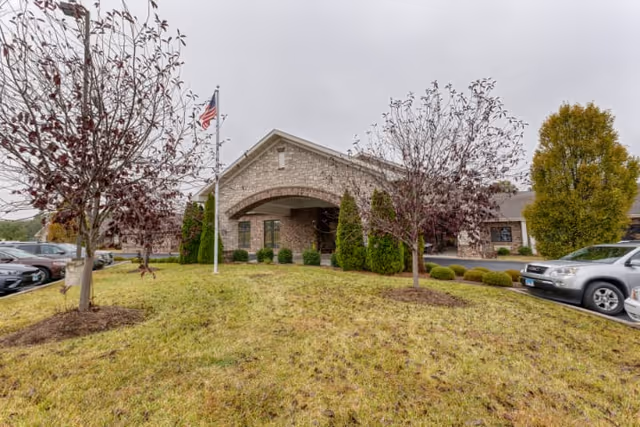 Exterior view of Cedarhurst Senior Living of Waterloo showing a single-story brick building with an arched entrance. There is a grassy area with two small trees and an American flag on a flagpole in front. Several parked cars are visible on either side of the building.