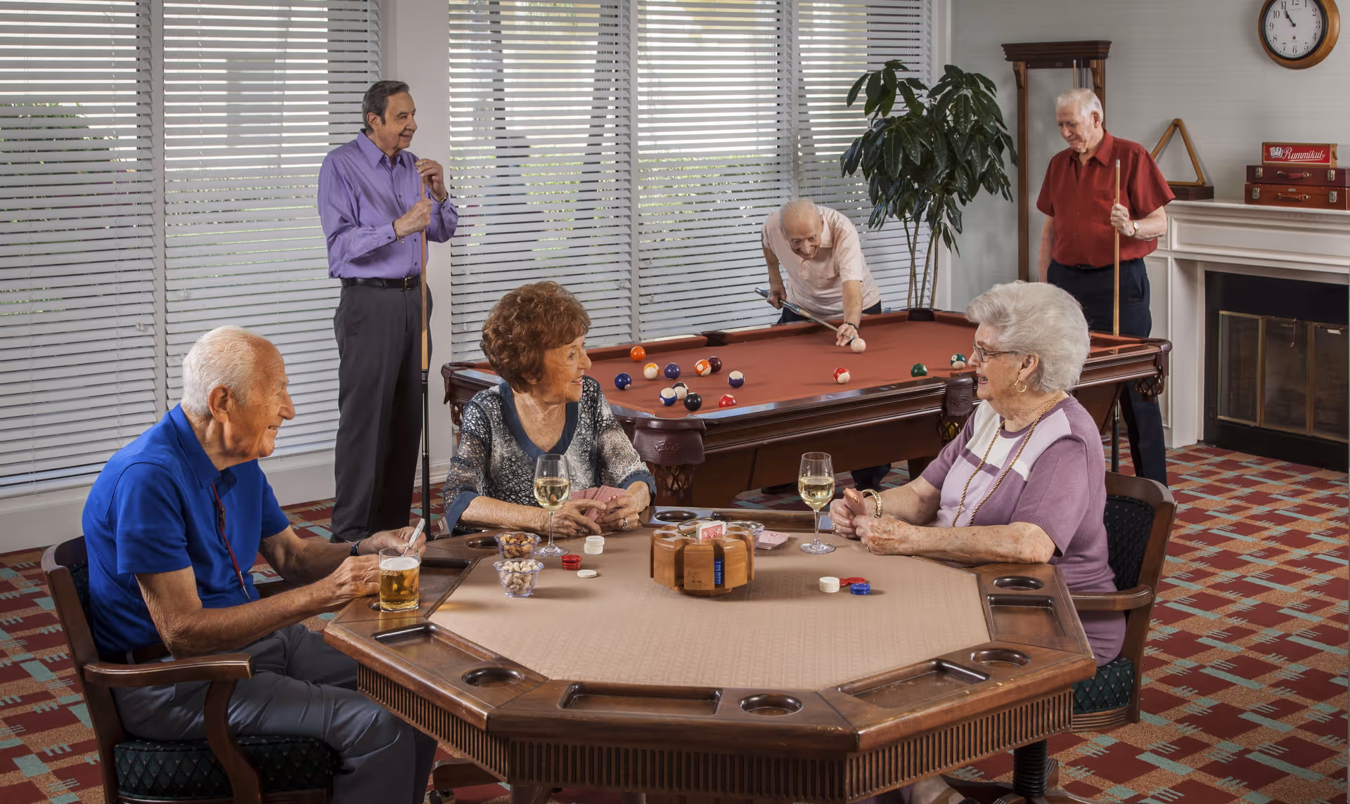 A group of elderly people enjoying leisure activities in a common room. Three seniors are seated around a card table with drinks and snacks, while two men are playing pool in the background. The room has large windows with blinds, a fireplace, and a patterned carpet.