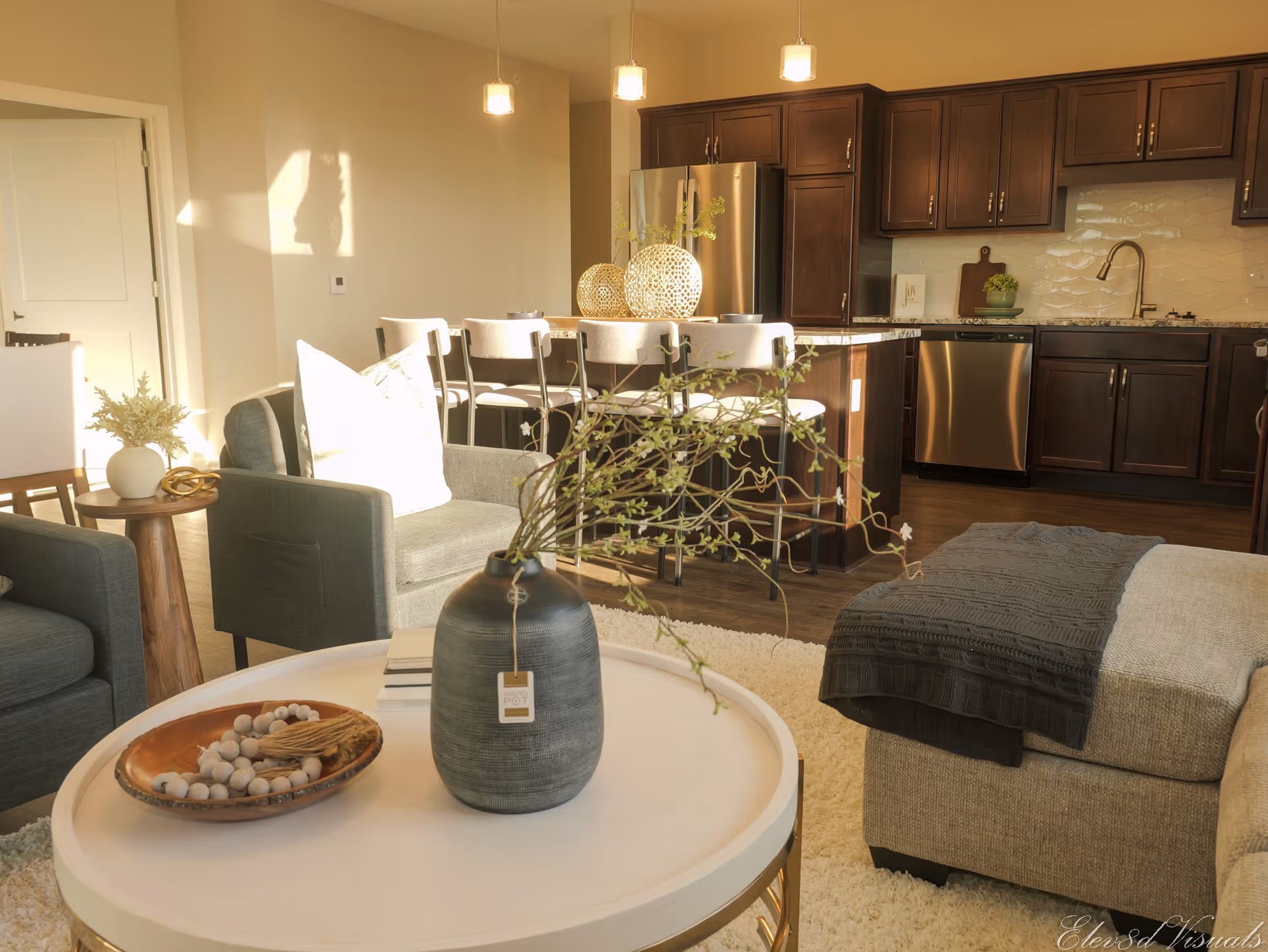 Open-plan living area with a round coffee table and vase in the foreground, upholstered seating and an island kitchen with dark cabinets in the background.
