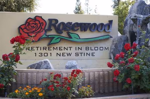 Exterior sign for Rosewood Retirement in Bloom, surrounded by red flowers.